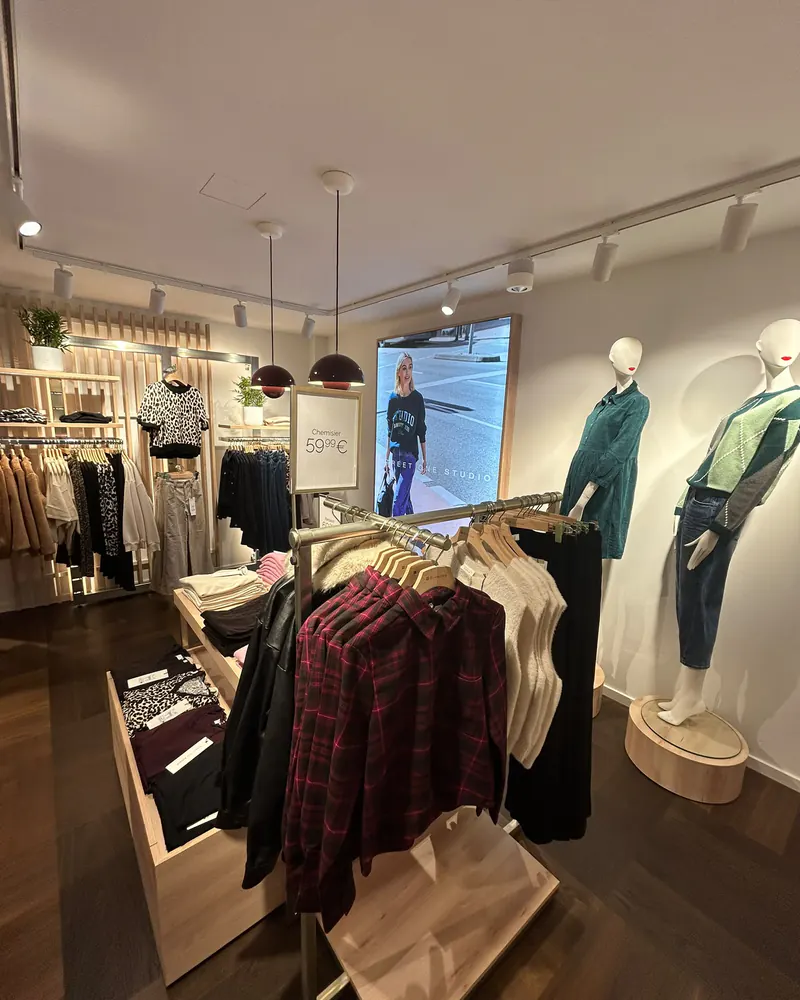 Le coin STREET ONE STUDIO : un portant de chemises à carreaux et de maille écrue, un grand écran et deux mannequins habillés de vert